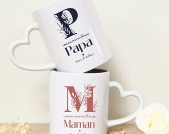 May include: Two white ceramic mugs with heart-shaped handles. The top mug has a black floral design with the letter "P" and the text "comme merveilleux Papa Clara et Arthur". The bottom mug has a brown floral design with the letter "M" and the text "comme merveilleuse Maman Clara et Arthur".