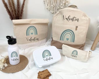 Puede incluir: Un conjunto de accesorios escolares o de viaje beige y blancos, que incluye una mochila, una bolsa de almuerzo, un estuche, una botella de agua y una fiambrera. Cada artículo presenta un diseño de arcoíris turquesa y el nombre "Valentin".