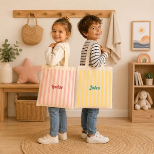 Peut inclure: Deux sacs fourre-tout rectangulaires en toile à rayures verticales. Un sac présente des rayures rose clair et le prénom Louise en écriture rouge. L'autre sac présente des rayures jaunes et le prénom Jules en écriture verte. Deux anses.