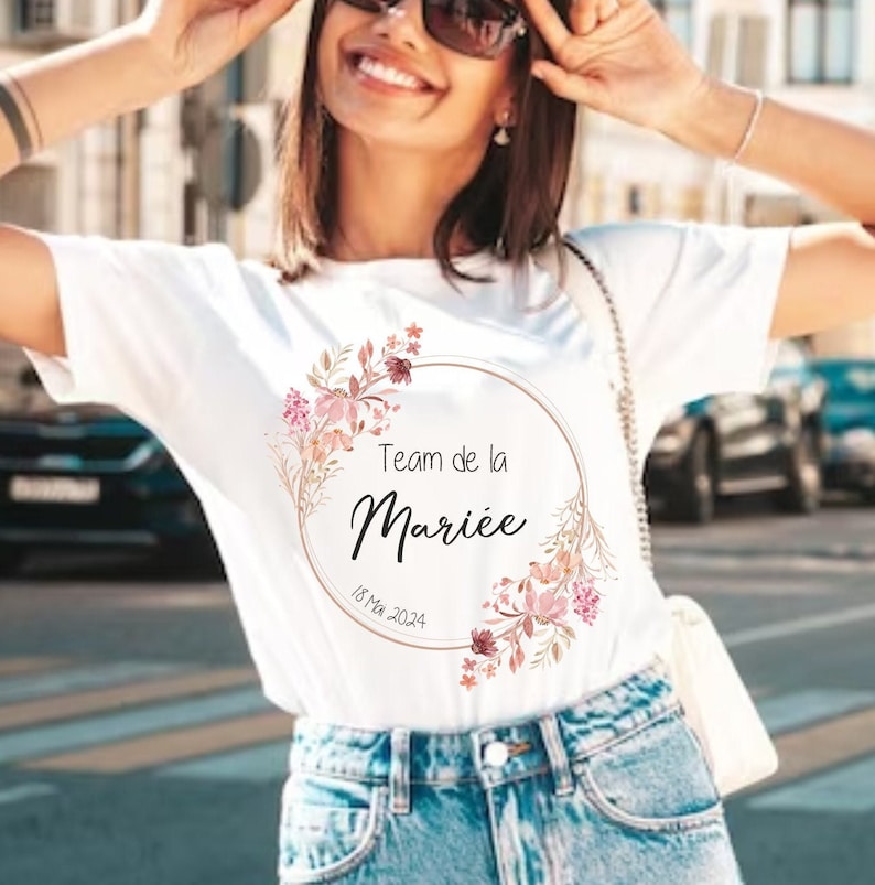 May include: White t-shirt with a floral design and the text "Team de la Mari&eacute;e" in black script, with the date "18 Mai 2024" below. The design is encircled by a gold-colored ring and floral accents in pink and brown.