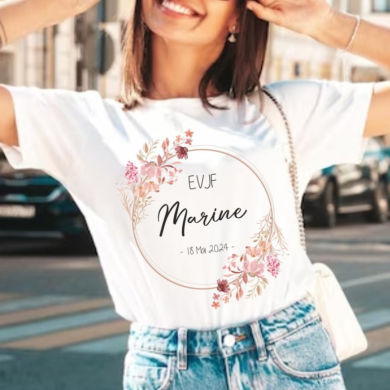 May include: White t-shirt with a floral wreath design. The design includes a circular frame with the name "Marine" in cursive, the initials "EVJF", and the date "18 Mai 2024". The wreath features pink and burgundy flowers.