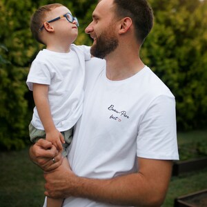 May include: A white t-shirt with the text "Beau-Pere fact" and a small red heart. A man is holding a young boy wearing a white t-shirt.