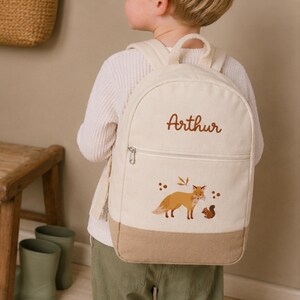 May include: A cream-coloured canvas backpack featuring a top handle and adjustable shoulder straps. The front displays the embroidered name Arthur in brown script, a zippered pocket, and a printed graphic of a fox and a squirrel in earthy tones.