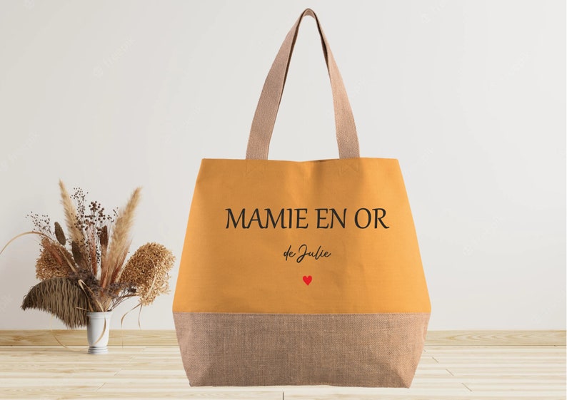 K&ouml;nnte beinhalten: Eine gelbe Tragetasche mit dem Text "MAMIE EN OR de Julie" und einem kleinen roten Herz. Die Tasche hat einen braunen Boden.