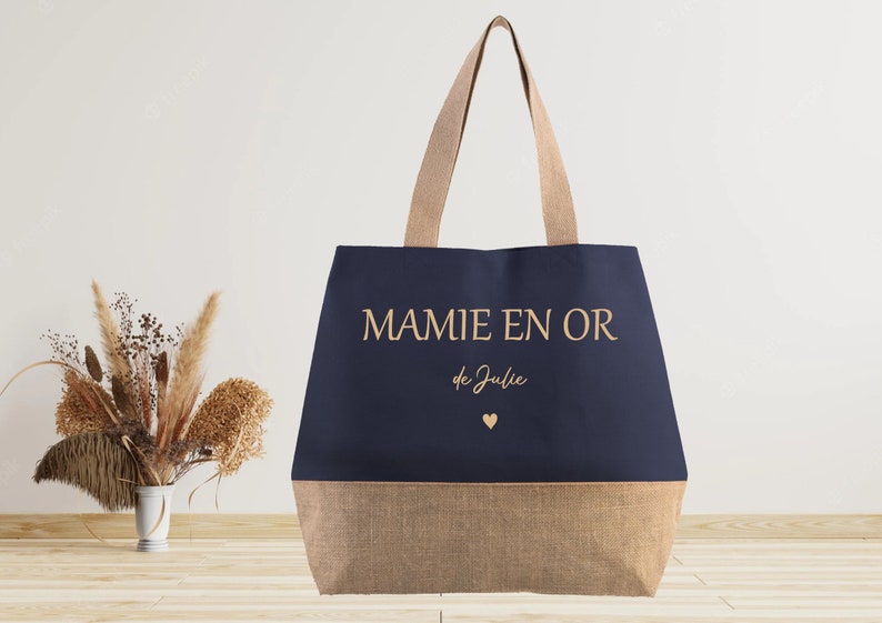 K&ouml;nnte beinhalten: Eine marineblaue Tragetasche mit braunem Juteboden. Die Tasche tr&auml;gt den Schriftzug "MAMIE EN OR de Julie" in goldenen Buchstaben mit einem kleinen goldenen Herz.