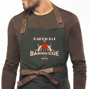 Peut inclure: Tablier vert foncé avec des lanières en cuir marron et une ceinture marron. Le tablier porte l'inscription "PAPI le Roi du BARBECUE Léon" en blanc, avec un dessin de flamme et de grill.