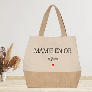 K&ouml;nnte beinhalten: Eine beige Tragetasche mit Juteboden. Die Tasche hat schwarzen Text, der "MAMIE EN OR de Julie" mit einem kleinen roten Herzen lautet.