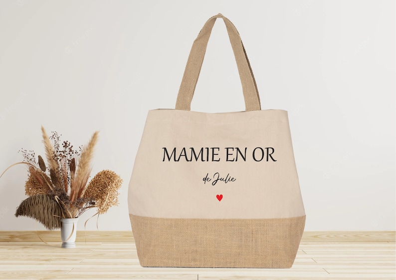K&ouml;nnte beinhalten: Eine beige Tragetasche mit Juteboden. Die Tasche hat schwarzen Text, der "MAMIE EN OR de Julie" mit einem kleinen roten Herzen lautet.