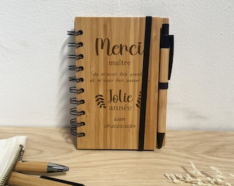 May include: A wooden notebook with a black spiral binding and a black elastic band closure. The notebook has a message engraved on the front: "Merci maître de m'avoir fait grand et m'avoir fait passer Jolie année Liam CP-2023/2024". A black and brown pen is attached to the notebook.