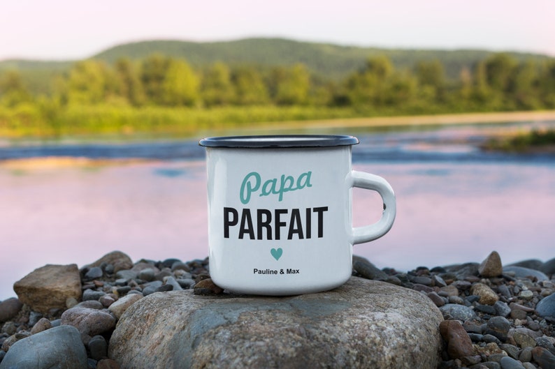 Pu&ograve; includere: Tazza in smalto bianco con bordo e manico neri. La tazza ha un design nero e turchese che dice "Papa Parfait" con un piccolo cuore turchese e il testo "Pauline & Max".