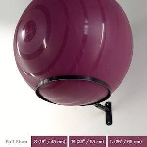 May include: A plum-coloured exercise ball is mounted on a black metal wall bracket. The ball features concentric circle designs. The image also shows the ball sizes: Small (45 cm), Medium (55 cm), and Large (65 cm).