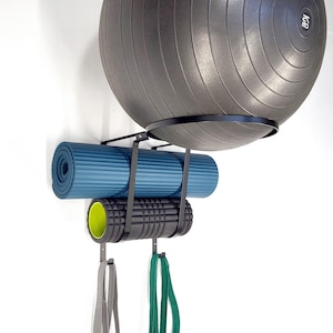 May include: A black wall-mounted fitness equipment organizer. It holds a gray exercise ball, a rolled-up blue yoga mat, a black foam roller with a yellow interior, and exercise bands. The organizer is mounted on a white wall.