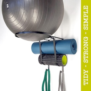 May include: A black metal wall-mounted organiser for fitness equipment. It holds a grey exercise ball, a blue yoga mat, a black foam roller, and resistance bands. The side text reads "TIDY - STRONG - SIMPLE."