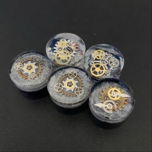 May include: Five clear resin plugs with embedded clockwork gears. The gears are a mix of gold and silver tones, set against a backdrop of silver glitter and dark blue swirls. The plugs are round and have a steampunk aesthetic.