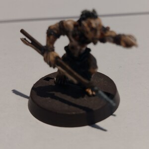 May include: A small, painted, plastic figurine of a goblin holding a spear. The goblin is painted in shades of brown, green, and grey. The figurine is standing on a black base.