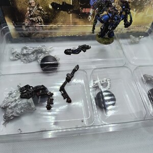 May include: A clear plastic tray with eight compartments containing miniature figures and accessories for a tabletop game. The figures are made of metal and painted in various colors, including silver, blue, and black. The accessories include weapons, bases, and other game pieces.