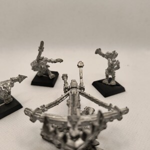 May include: A set of four miniature metal figures, including a catapult, two orcs with spears, and one orc with a sword. The figures are painted in a silver color.