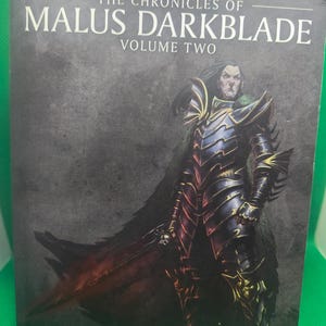 May include: The cover of a fantasy novel titled "The Chronicles of Malus Darkblade, Volume Two". The cover features a dark-haired man in black armor with a sword, standing against a dark gray background.