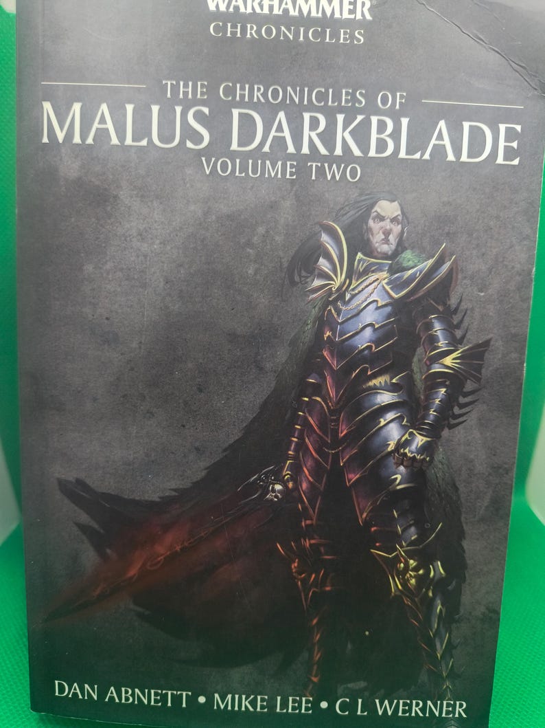 May include: The cover of a fantasy novel titled "The Chronicles of Malus Darkblade, Volume Two". The cover features a dark-haired man in black armor with a sword, standing against a dark gray background.