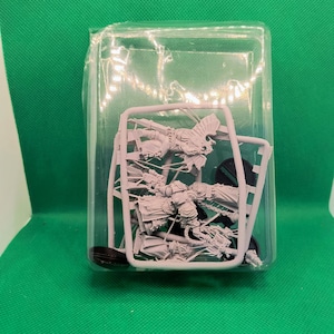 May include: A miniature model kit, still on its plastic sprues, encased in a clear plastic package. The model appears to be a detailed figure, possibly for tabletop gaming or hobby projects. The background is a solid green.