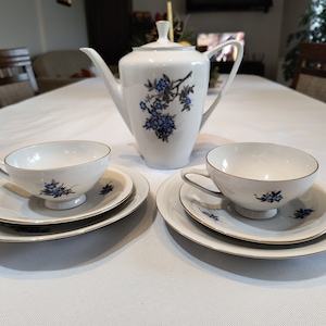 Może przedstawiać: Biały porcelanowy zestaw do herbaty z niebieskimi wzorami kwiatowymi. Zestaw zawiera czajnik, dwie filiżanki i dwa spodeczki.