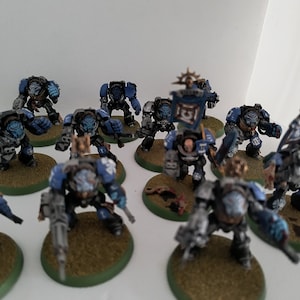 May include: A group of blue and silver painted miniature space marine figures, standing on round bases. The figures are holding weapons and some have flags.