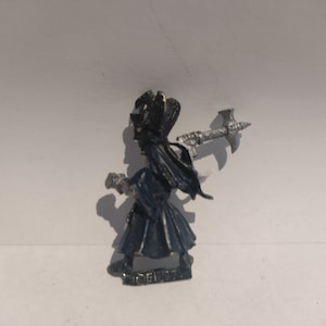 May include: A black and silver miniature figure of a person wearing a hooded robe and holding a weapon. The figure is standing on a small base.