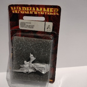 May include: A silver metal miniature of a female figure in a flowing robe, holding a sword. The miniature is packaged in a plastic blister pack with a red and black card backing that reads "Warhammer" and "Undead Banshee".