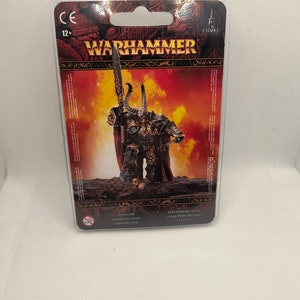 May include: A plastic blister pack containing a miniature plastic figure of a Chaos Lord from the Warhammer 40,000 universe. The figure is painted in black and red armor with a large sword and a skull on its base. The packaging is red and black with the Warhammer logo and the text "Chaos Lord" and "Kriegsherr des Chaos".