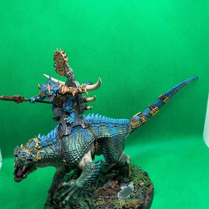May include: A blue and green miniature figure of a lizard-like creature riding a larger lizard-like creature. The smaller creature is holding a spear and has a golden crown on its head. The larger creature has a long tail and is standing on a base with green foliage.