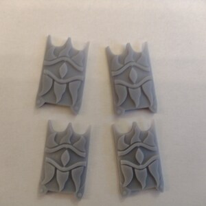 Puede incluir: Cuatro piezas de plástico gris con un diseño decorativo. Cada pieza tiene un patrón repetitivo de un ojo estilizado, una línea ondulada y una serie de formas puntiagudas.