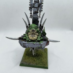 May include: A green and white miniature figure of a goblin sitting on a throne with a spiked base. The throne is decorated with a stone structure and a banner. The goblin is holding a staff and has a large belly.