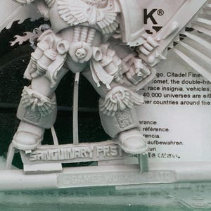 May include: A gray plastic miniature figure of a warrior in detailed armor, with a stern expression and raised weapon. The figure is labeled "SANGUINARY PRST" and "© GAMES WORKSHOP LTD". The figure is part of a tabletop game.