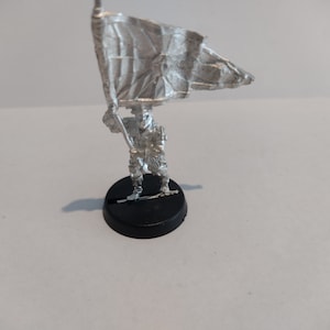 May include: A silver miniature figure of a soldier holding a flag. The flag is white with a black design. The figure is standing on a black base.