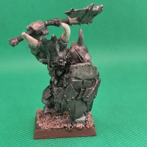 May include: A miniature figure of a heavily armored, horned, and muscular warrior holding a large axe. The figure is painted in shades of gray and black, with a brown base.