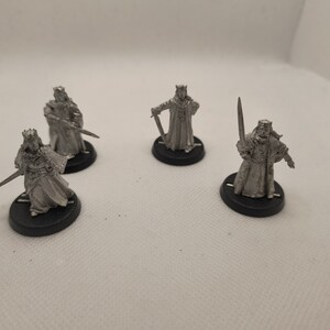 May include: Four silver-colored miniature figures of a king wearing a crown and robe, each holding a sword. The figures are standing on black circular bases.