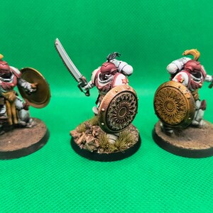 May include: Three white and red painted miniature figures with gold shields and swords. The figures are standing on brown bases.