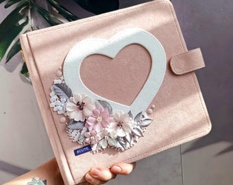 May include: A pink suede album with a white heart-shaped cutout and a floral embellishment. The album has a gold zipper and a suede strap. The text "#CUTE" is printed on the front of the album.