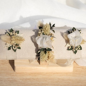 Peut inclure: Présentoir rectangulaire beige avec trois compositions florales. Chaque composition présente des fleurs séchées, de la verdure et des accents blancs. Le présentoir est sur une base en bois clair, avec un fond en tissu blanc.
