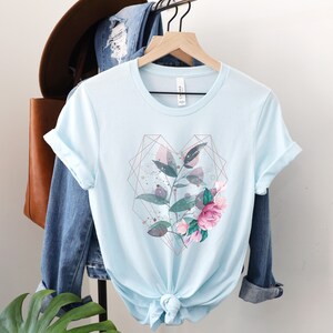 Puede incluir: Una camiseta azul claro con un dise&ntilde;o floral en forma de coraz&oacute;n geom&eacute;trico. El dise&ntilde;o presenta flores y hojas rosas y verdes con detalles dorados.