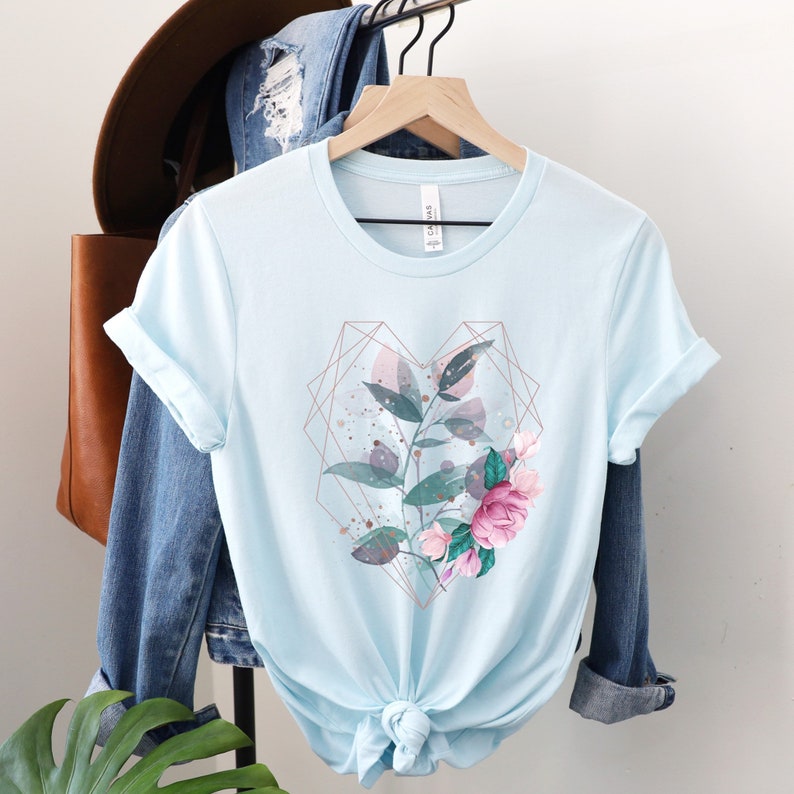 Puede incluir: Una camiseta azul claro con un dise&ntilde;o floral en forma de coraz&oacute;n geom&eacute;trico. El dise&ntilde;o presenta flores y hojas rosas y verdes con detalles dorados.