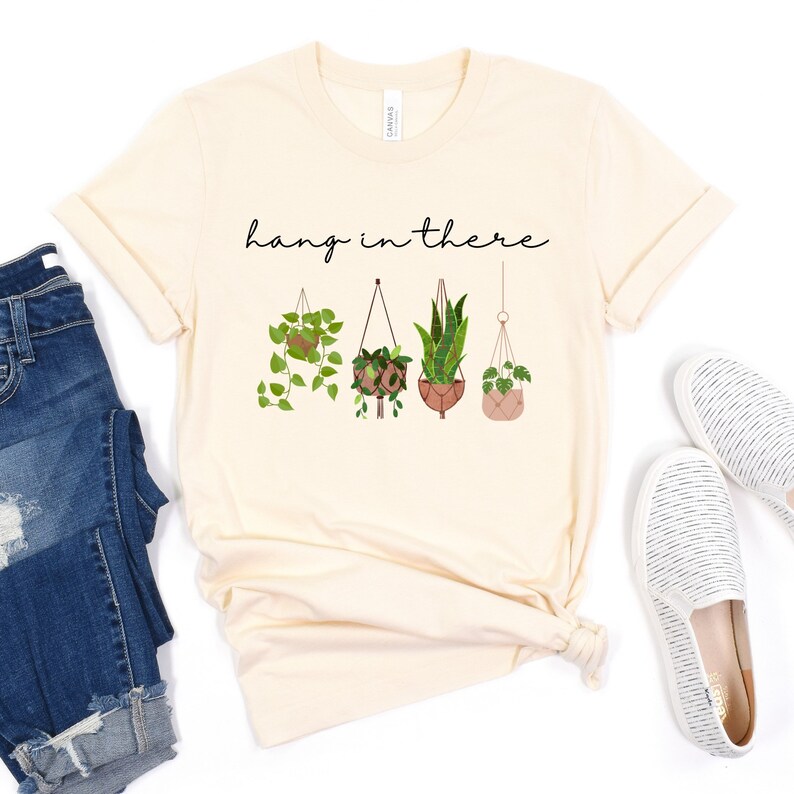 May include: Cream-colored t-shirt featuring the phrase "hang in there" in black script above four hanging potted plants. The plants are in various styles of hanging planters, with green leaves and brown pots. The shirt is styled with blue jeans and white slip-on shoes.