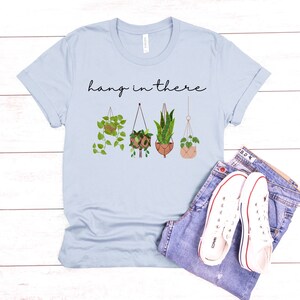 May include: Light blue t-shirt with the phrase "hang in there" in black script. The shirt features a graphic of four hanging plants in various pots. A pair of blue jeans and white sneakers complete the casual look.
