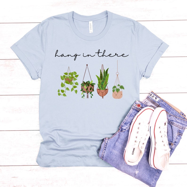 May include: Light blue t-shirt with the phrase "hang in there" in black script. The shirt features a graphic of four hanging plants in various pots. A pair of blue jeans and white sneakers complete the casual look.
