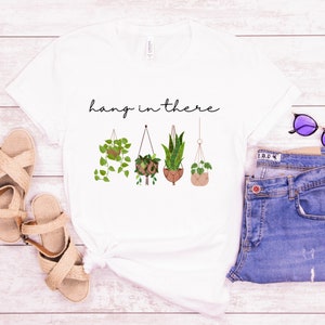 May include: White t-shirt featuring a botanical design of four hanging plants in pots. The phrase "hang in there" is written above the plants in a cursive font. The shirt is paired with blue jeans and sandals.