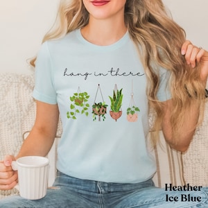 May include: Heather Ice Blue t-shirt with the phrase "hang in there" in black script. Below the text are illustrations of four hanging plants in various pots. The shirt is a light blue color.