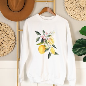 Peut inclure: Sweat-shirt blanc avec un motif d'arbre de citron. Le motif représente une branche de citronnier avec des feuilles vertes, des fleurs blanches et des citrons jaunes.
