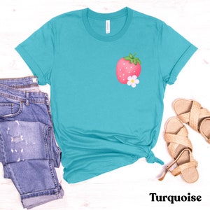 May include: A turquoise t-shirt with a pink strawberry and white flower graphic. The shirt is laid flat on a white wooden surface with a pair of brown sandals and a pair of blue jeans.