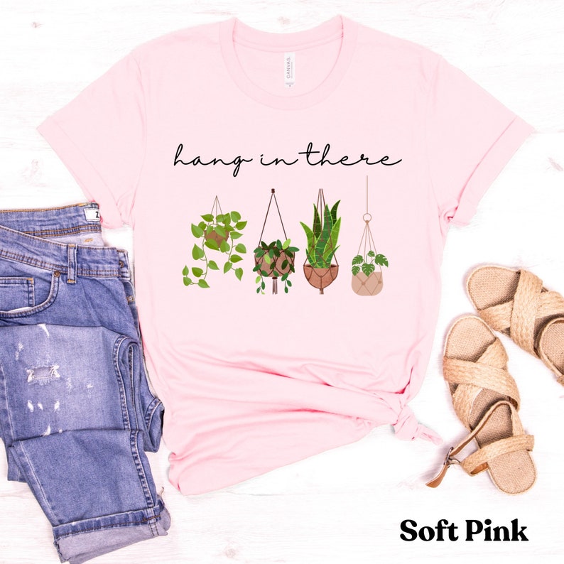 May include: Soft pink t-shirt with the phrase "hang in there" above a graphic of four hanging plants. The plants are in various pots and macrame hangers. The shirt is displayed with blue jeans and sandals.