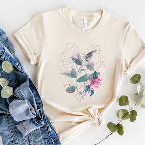 Puede incluir: Una camiseta de color crema con un dise&ntilde;o floral dentro de una forma de coraz&oacute;n geom&eacute;trico. El dise&ntilde;o presenta flores y hojas de acuarela rosa y verde.